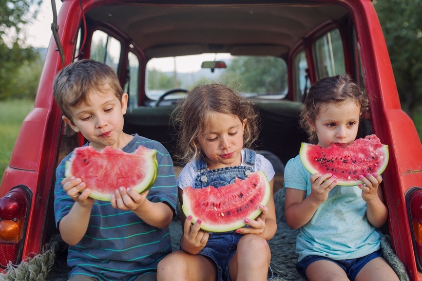 two young Girls and a boy eating watermelon on back of car