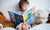 boy sitting on a bed reading story book surrounded with toys