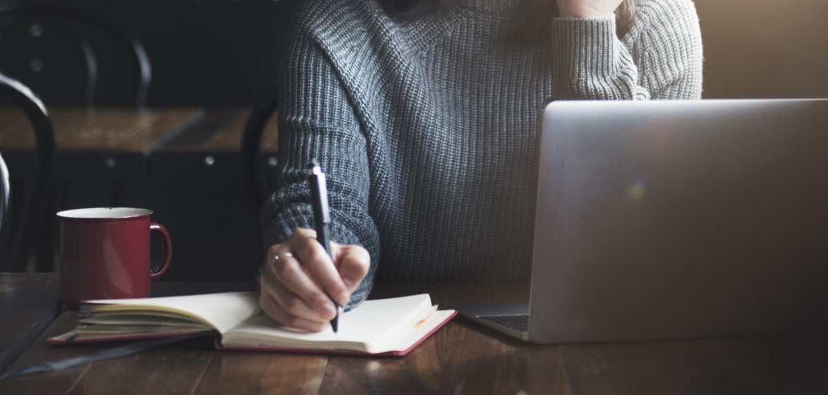 A Person writing something in a book and a laptop opened